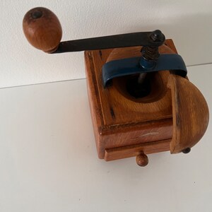 Puede incluir: Un molinillo de caf&eacute; cuadrado de madera con una manivela de metal oscuro con un pomo de madera redondo, un mecanismo de puente de metal azul y un peque&ntilde;o caj&oacute;n extra&iacute;ble con un tirador de madera diminuto. La tapa superior est&aacute; entreabierta.