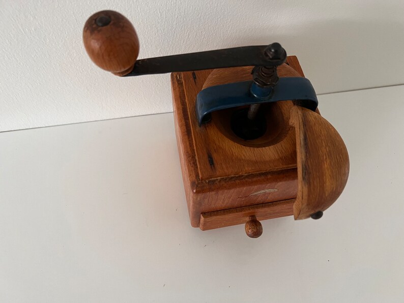 Puede incluir: Un molinillo de caf&eacute; cuadrado de madera con una manivela de metal oscuro con un pomo de madera redondo, un mecanismo de puente de metal azul y un peque&ntilde;o caj&oacute;n extra&iacute;ble con un tirador de madera diminuto. La tapa superior est&aacute; entreabierta.
