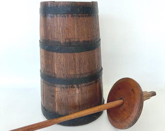 May include: A wooden butter churn with a wooden paddle. The churn is made of wood staves and has three metal bands around it. The paddle has a round wooden handle and a flat wooden blade.