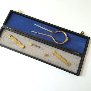 May include: A black rectangular case with a blue interior containing a brass divider tool and a clear plastic drafting ruler. The ruler features brass knobs and the text "W.H. Harling" and "4742" printed on its surface. The set is for technical drawing.