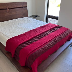 May include: A wooden bed frame with a white mattress and a red and brown striped blanket. The bed is in a modern bedroom with a white wall and a window.