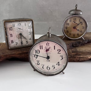 Puede incluir: Tres relojes despertadores vintage con cajas de metal. Los relojes tienen caras blancas con números y manecillas negras. El reloj de la izquierda tiene una caja cuadrada, el reloj del centro tiene una caja redonda y el reloj de la derecha tiene una caja redonda con una campana en la parte superior.