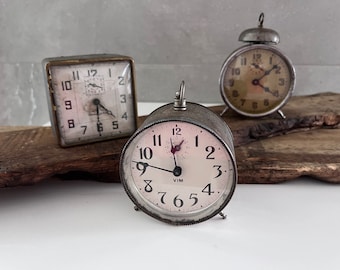 May include: Three vintage alarm clocks with metal cases. The clocks have white faces with black numbers and hands. The clock on the left has a square case, the clock in the center has a round case, and the clock on the right has a round case with a bell on top.