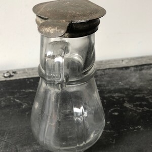 May include: A clear glass pitcher with a metal lid. The pitcher has a rounded body and a handle. The lid is slightly tarnished.