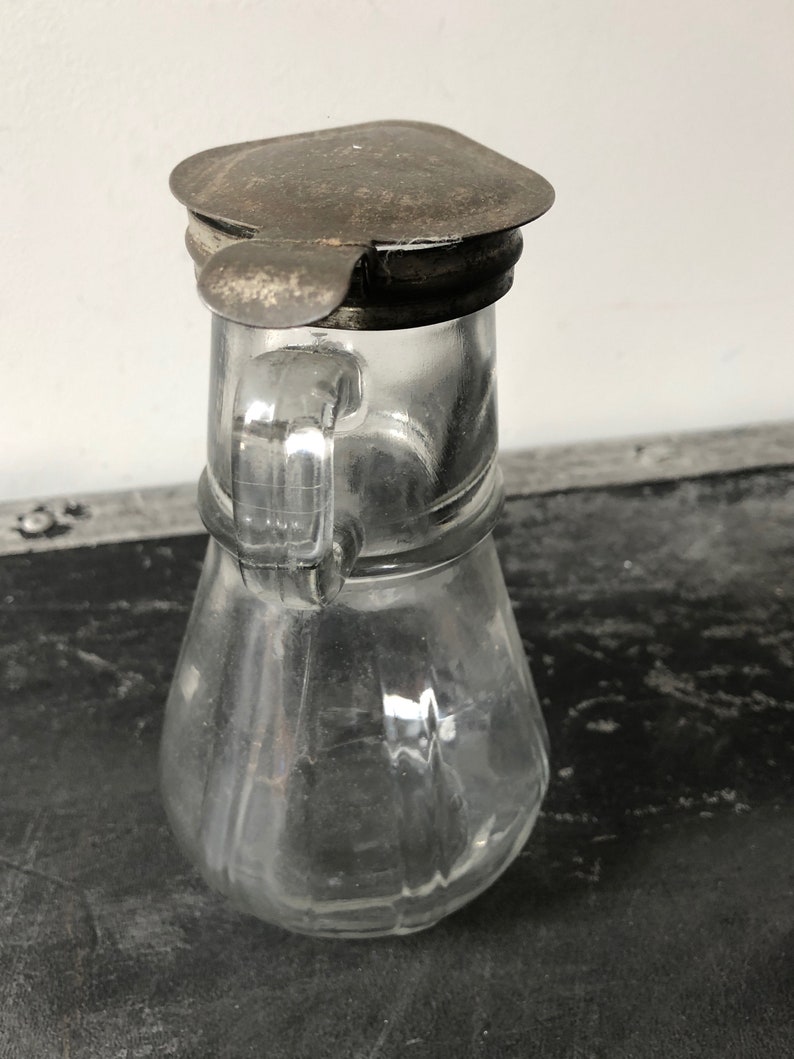 May include: A clear glass pitcher with a metal lid. The pitcher has a rounded body and a handle. The lid is slightly tarnished.