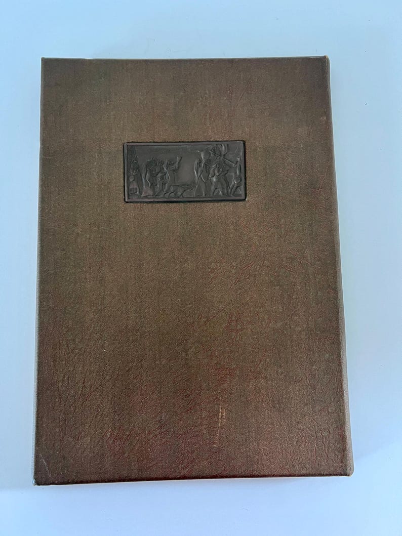 Puede incluir: Un libro encuadernado en cuero marr&oacute;n con una imagen en relieve de una escena con varias figuras en la portada.