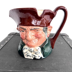 May include: A novelty ceramic jug in the shape of a man's head, with a winking face. It has a dark brown hat, a green jacket, and a white cravat with blue spots. The handle is dark brown. A decorative item.