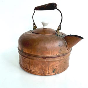May include: A vintage copper kettle with a dark wooden handle and a white ceramic knob on the lid. The kettle has a spout and a curved metal handle. The copper has a patina, showing its age and use.