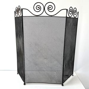May include: Black metal fireplace screen with a decorative scrolled top. The screen has a mesh design and is supported by small, round feet. The top features three scrolled accents.