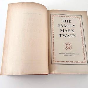 Puede incluir: Un libro de tapa dura abierto titulado The Family Mark Twain, publicado por Harper & Brothers Publishers en Nueva York y Londres. La página del título presenta un borde decorativo rojo y un emblema circular, con texto negro sobre una página color crema.