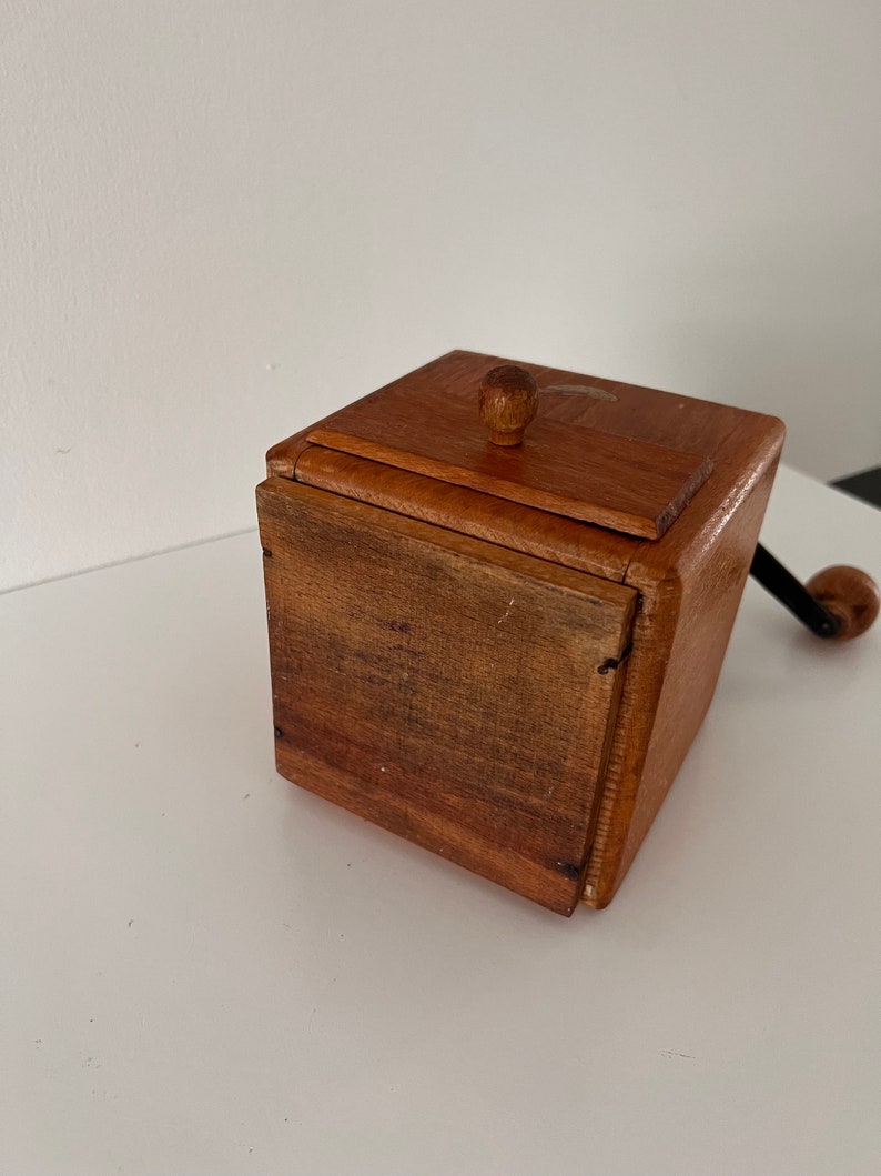 Puede incluir: Un molinillo de caf&eacute; cuadrado de madera con acabado marr&oacute;n medio, que presenta una tapa superior con bisagras y un peque&ntilde;o pomo redondo de madera, un caj&oacute;n frontal con veta de madera oscura y una manivela de metal negro con un pomo de madera.