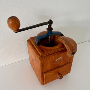 Puede incluir: Un molinillo de caf&eacute; cuadrado de madera con acabado marr&oacute;n claro, manivela de metal oscuro con pomo de madera, soporte superior de metal azul y un peque&ntilde;o caj&oacute;n extra&iacute;ble con tirador redondo de madera. Tiene una peque&ntilde;a calcoman&iacute;a al frente.