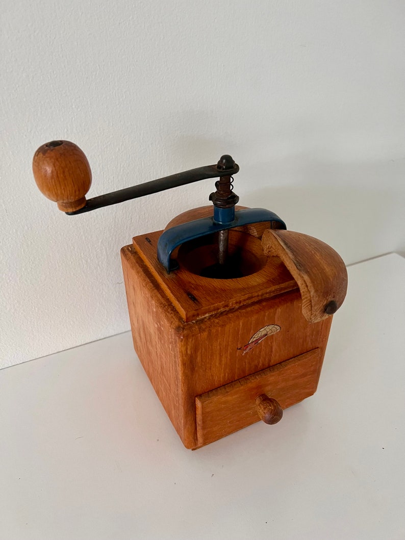Puede incluir: Un molinillo de caf&eacute; cuadrado de madera con acabado marr&oacute;n claro, manivela de metal oscuro con pomo de madera, soporte superior de metal azul y un peque&ntilde;o caj&oacute;n extra&iacute;ble con tirador redondo de madera. Tiene una peque&ntilde;a calcoman&iacute;a al frente.