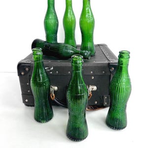 May include: A collection of vintage green glass bottles, arranged on a dark suitcase. The bottles have a classic soda bottle shape with vertical ribbing. The bottles are empty and have a glossy finish.