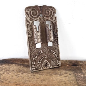 May include: An antique, cast iron clip with an ornate, scrollwork design. The clip has two slots and the text "THE GURNEY FOUNDRY CO. TORONTO" is embossed on the front. The clip is a weathered, metallic color.