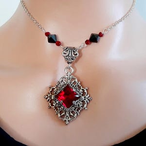 May include: Silver-toned pendant necklace with a large, square, red gemstone. The pendant is ornate with a filigree design. The necklace has a delicate chain with small red and black accents. The Alchemist's Cauldron is printed at the bottom right.