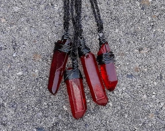 May include: Four red crystal pendants suspended from black metal chains. Each translucent red stone is wrapped in dark wire at the top. The text THE ALCHEMIST'S CAULDRON is visible at the bottom left of the image.