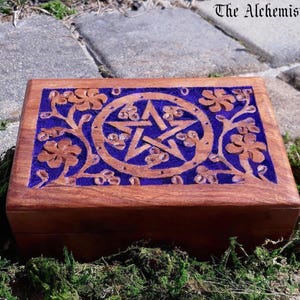 May include: A rectangular wooden box with a hinged lid featuring a carved pentagram inside a circle, surrounded by floral vine patterns. The carved areas are filled with a vibrant purple inlay. The text The Alchemist's Cauldron is visible.