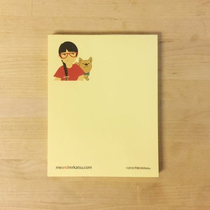 May include: A yellow notepad with a cartoon illustration of a girl with red glasses and a dog. The notepad has the text "meandmrkatsu.com" and "©2018 meandmrkatsu" printed on the bottom.