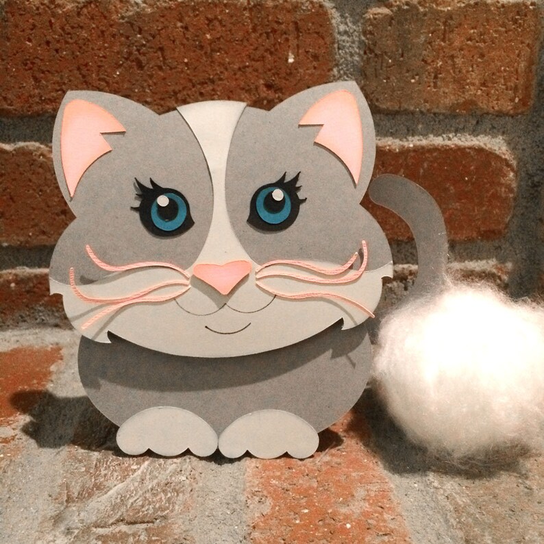 Op de afbeelding: Een grijze papieren kat met roze oren en een witte, pluche staart. De kat heeft blauwe ogen en een roze neus.