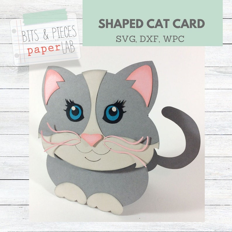 Op de afbeelding: Een grijze en roze papieren knutselkattenkaart met een wit gezicht en blauwe ogen. De kaart is katvormig en heeft een staart die naar de zijkant uitsteekt. De kaart is gelabeld met "Shaped Cat Card" en "SVG, DXF, WPC".
