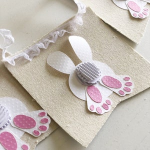 May include: A white fabric banner with a white bunny tail and pink footprints. The bunny tail is made of paper and has a white polka dot pattern. The banner is decorated with white lace.