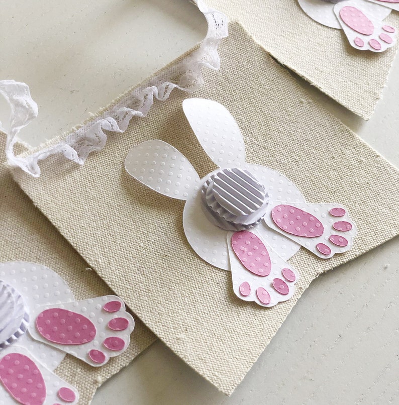 May include: A white fabric banner with a white bunny tail and pink footprints. The bunny tail is made of paper and has a white polka dot pattern. The banner is decorated with white lace.