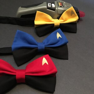 May include: Three bow ties with black and colored fabric. The top bow tie is yellow with a gold star trek insignia. The middle bow tie is blue with a gold star trek insignia. The bottom bow tie is red with a gold star trek insignia.
