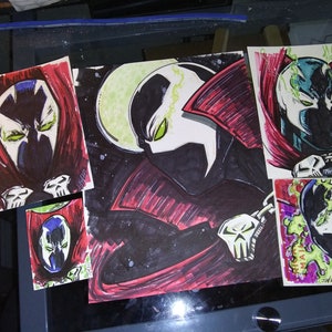 May include: Four black and white drawings of the comic book character Spawn. The drawings are done in black marker and colored with red, green, and yellow markers. The drawings are on white paper.