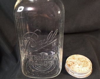 Peut inclure: Un bocal en verre transparent Ball avec un couvercle blanc. Le bocal a une surface texturée et les mots "Ball" et "Perfect Mason" sont en relief sur le devant.