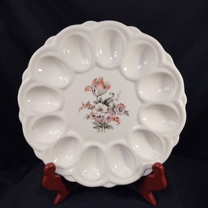 May include: White ceramic egg plate with 12 individual indentations for serving eggs. The plate features a floral design with pink and orange flowers.