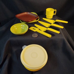 May include: A set of yellow plastic kitchenware, including a bowl, a plate, a cup, a lidded container, and four butter knives. The items are arranged on a black surface.