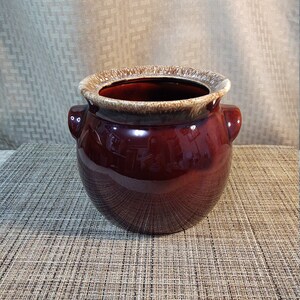 Puede incluir: Una olla de cerámica marrón con un borde glaseado marrón y blanco. La olla tiene dos pequeñas asas a los lados.