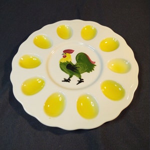 May include: White ceramic egg plate with a scalloped edge. The plate has 10 yellow egg-shaped indentations and a painted rooster in the center.