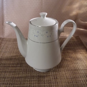 May include: White ceramic coffee pot with a floral design and a silver trim. The pot has a lid and a handle.
