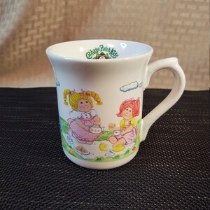 Op de afbeelding: Een witte keramische mok met een groen "Cabbage Patch Kids" logo en een cartoon afbeelding van twee poppen die een picknick houden. De poppen dragen roze en gele jurken en zitten op een groene deken met een picknickmand, een theepot en koekjes.
