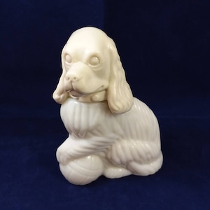 Puede incluir: Figura de cerámica blanca de un cocker spaniel sentado con la cabeza inclinada hacia un lado. El perro lleva un collar y tiene una pelota a sus pies.