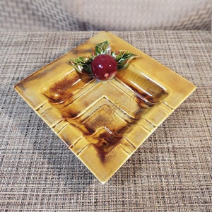 May include: A square ceramic ashtray with a brown and yellow glaze. The ashtray has a raised design of a red apple with green leaves.