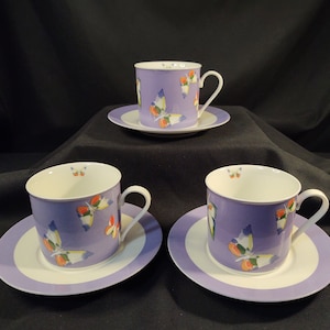 May include: Three purple and white teacups and saucers with a butterfly design. The cups and saucers have a white interior and a purple exterior with a white band around the rim. The butterflies are colorful and have orange, yellow, and blue accents.