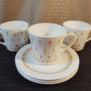 May include: Three white ceramic coffee mugs with a floral pattern of pink, yellow, and green tulips. The mugs are on a white saucer with a matching floral pattern.