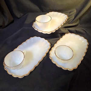 May include: Three white teacups and saucers with gold trim. Each saucer has a scalloped edge. The teacups are placed on the saucers. The set is on a dark background.