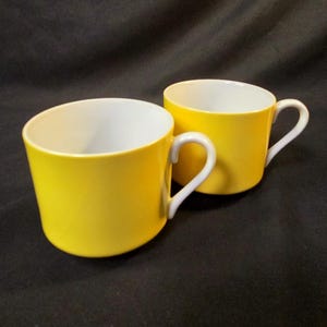 May include: Two yellow ceramic mugs with white interiors and white handles are displayed against a dark background. The mugs are side by side, showcasing their bright color and simple design. The handles are curved and easy to grip.