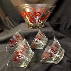 May include: A set of clear glass popcorn bowls, one large and four smaller. Each bowl has the words "Pop Corn" printed in red. The bowls have a flared design.