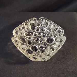 Peut inclure: Un cendrier carré en verre transparent avec une surface texturée. Le cendrier a un bord surélevé et un motif de petites bulles.