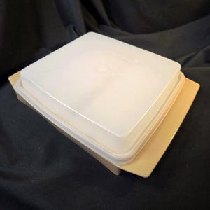 Könnte beinhalten: Ein quadratischer, cremefarbener Tupperware-Behälter mit passendem Deckel. Der Deckel ist durchscheinend und die Basis hat eine hellbraune Farbe. Der Behälter ist vor einem dunklen Hintergrund platziert.