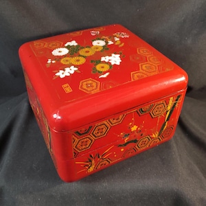 Può includere: Una scatola di legno laccata di rosso con un coperchio incernierato. La scatola è decorata con un motivo floreale in oro, nero e bianco. Il coperchio presenta un disegno floreale con crisantemi e altri fiori. La scatola è in stile giapponese tradizionale.