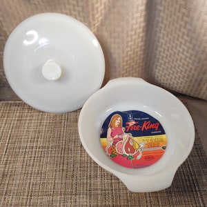 May include: A white Fire-King casserole dish with a lid. The dish is round with two handles and has a blue and red label that says "Fire-King Ovenware". The label features a woman holding a casserole dish and a graphic of a pineapple, ham, and pear.