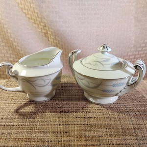 Peut inclure: Un ensemble de crémier et sucrier blanc avec un délicat motif de blé bleu et gris. Le crémier a une poignée et le sucrier a un couvercle.