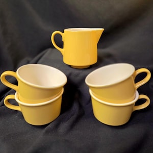 May include: A set of yellow ceramic cups and a creamer. The cups are stacked in pairs, with a white interior and a curved rim. The creamer has a handle and a spout. The items are arranged on a dark surface.