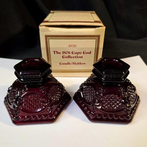 May include: Two ruby red glass candle holders from the Avon 1876 Cape Cod Collection. The candle holders have a hexagonal base with ornate detailing and a removable, faceted lid. The original box is in the background.